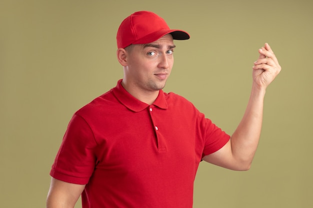 Foto gratuita repartidor joven complacido con uniforme y gorra mostrando gesto de punta aislado en la pared verde oliva