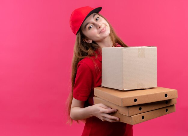 Repartidor joven complacido en uniforme rojo con caja y paquetes en espacio rosa aislado con espacio de copia