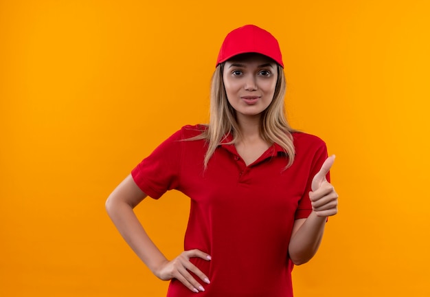 Repartidor joven complacido con uniforme rojo y gorra poniendo la mano en la cadera con el pulgar hacia arriba aislado sobre fondo naranja