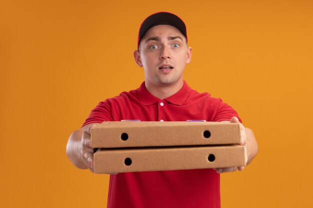 Repartidor joven complacido vestido con uniforme con gorra sosteniendo cajas de pizza en la parte delantera aislada en la pared naranja