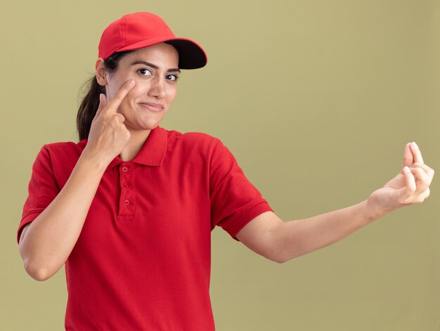 Repartidor joven complacido vistiendo uniforme con gorra poniendo el dedo en el ojo mostrando el gesto de la punta aislado en la pared verde oliva