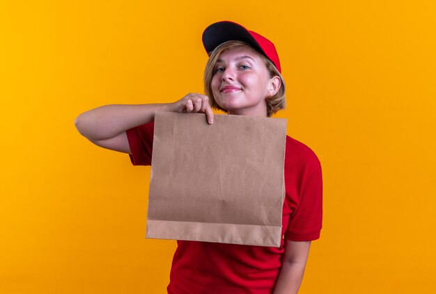 Repartidor joven complacido vistiendo uniforme con gorra sosteniendo una bolsa de comida de papel aislado en la pared naranja