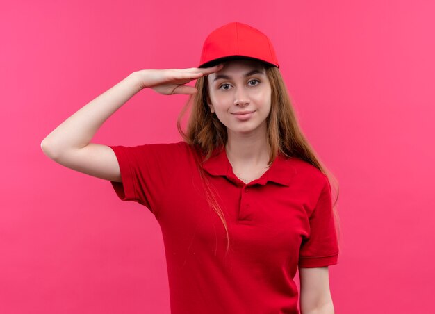 Repartidor joven confiado en uniforme rojo que pone la mano cerca de la frente en el espacio rosado aislado