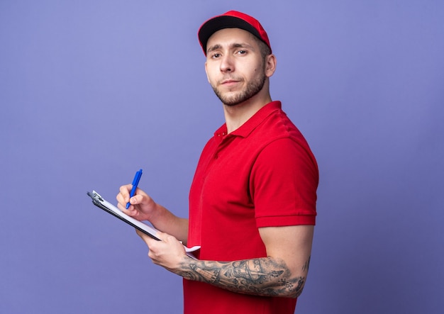 Foto gratuita repartidor joven confiado vistiendo uniforme con gorra escribiendo algo en el portapapeles