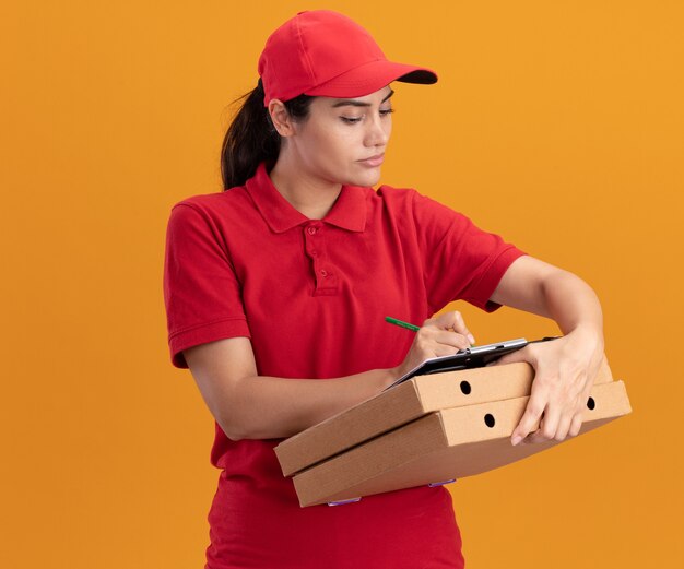 Repartidor joven confidente con uniforme y gorra escribiendo algo en el portapapeles en cajas de pizza aisladas en la pared naranja