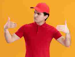 Foto gratuita repartidor joven confundido con uniforme rojo y gorra finge sostener algo frente a él mirando su mano aislada en la pared naranja