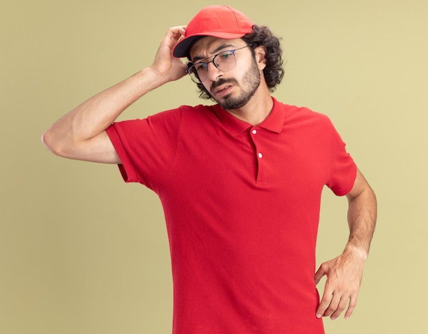 Repartidor joven confundido en uniforme rojo y gorra con gafas tocando la cabeza mirando hacia abajo aislado en la pared verde oliva