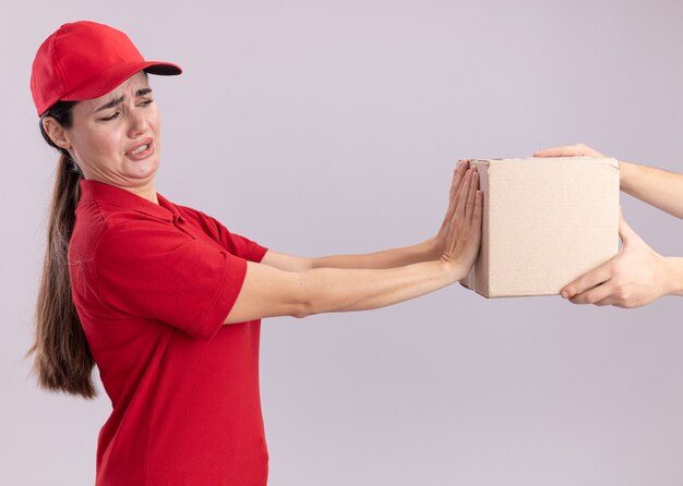 Repartidor joven descontento en uniforme y gorra de pie en la vista de perfil dando cardbox al cliente mirando la caja haciendo gesto de rechazo aislado en la pared blanca