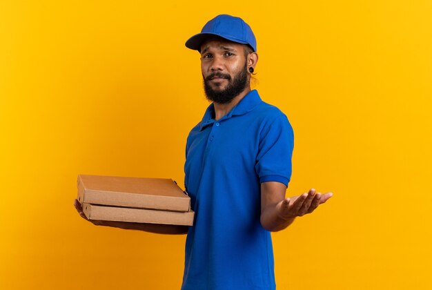 Repartidor joven despistado sosteniendo cajas de pizza y apuntando al frente aislado en la pared naranja con espacio de copia