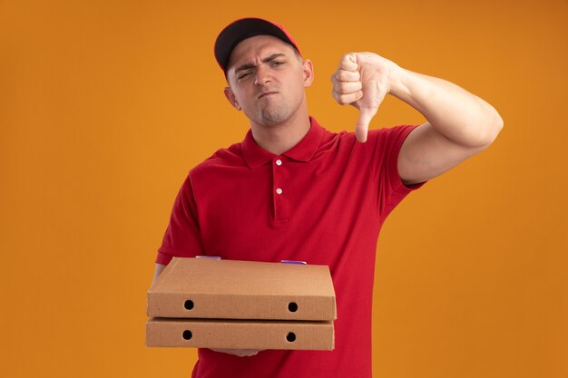 Repartidor joven disgustado con uniforme con gorra sosteniendo cajas de pizza mostrando el pulgar hacia abajo aislado en la pared naranja