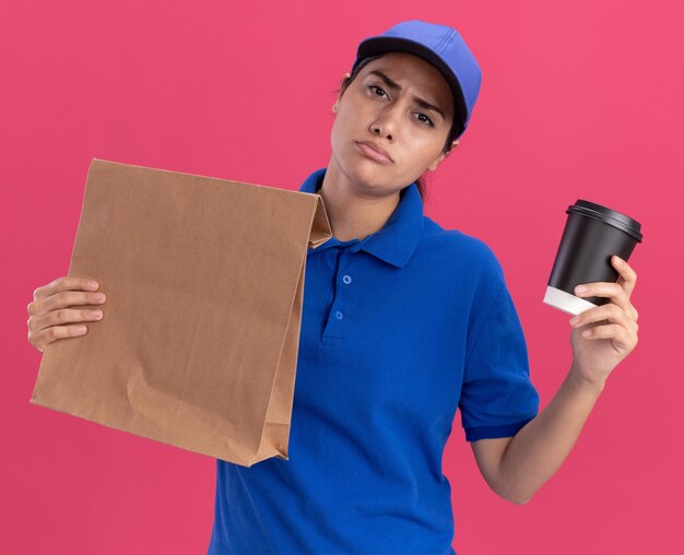 Repartidor joven disgustado con uniforme con gorra sosteniendo el paquete de alimentos de papel con una taza de café aislado en la pared rosa