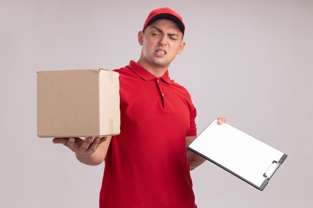 Repartidor joven disgustado con uniforme con gorra sosteniendo el portapapeles mirando la caja en la mano aislada en la pared blanca