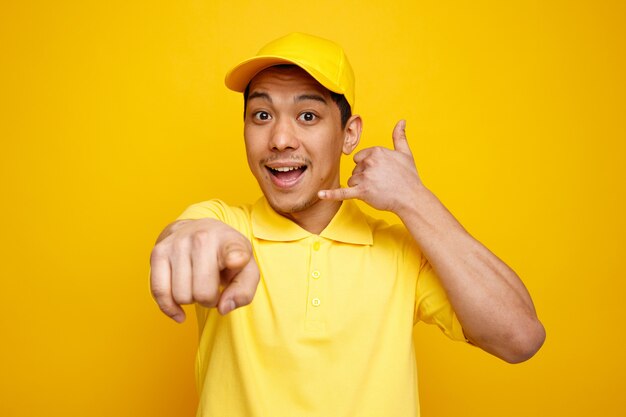 Repartidor joven emocionado con gorra y uniforme mirando y apuntando a la cámara haciendo gesto de llamada