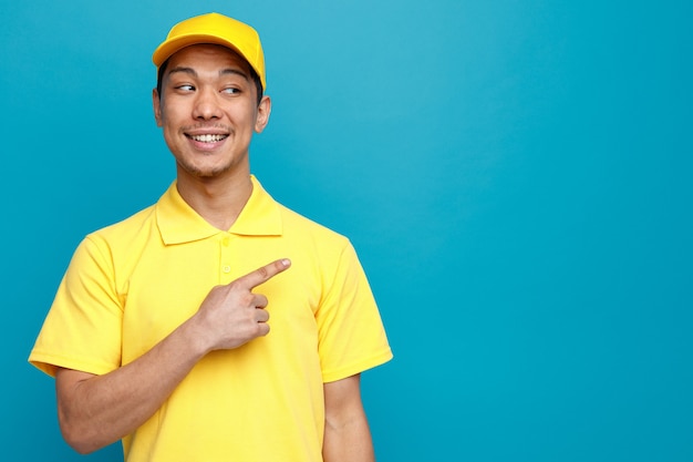 Repartidor joven emocionado vestido con uniforme y gorra mirando y apuntando a un lado