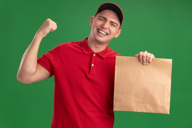 Repartidor joven emocionado vestido con uniforme y gorra sosteniendo el paquete de comida de papel que muestra sí gesto aislado en la pared verde