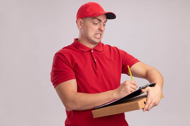 Repartidor joven enojado con uniforme con gorra sosteniendo la caja de pizza y escribiendo algo en el portapapeles aislado en la pared blanca