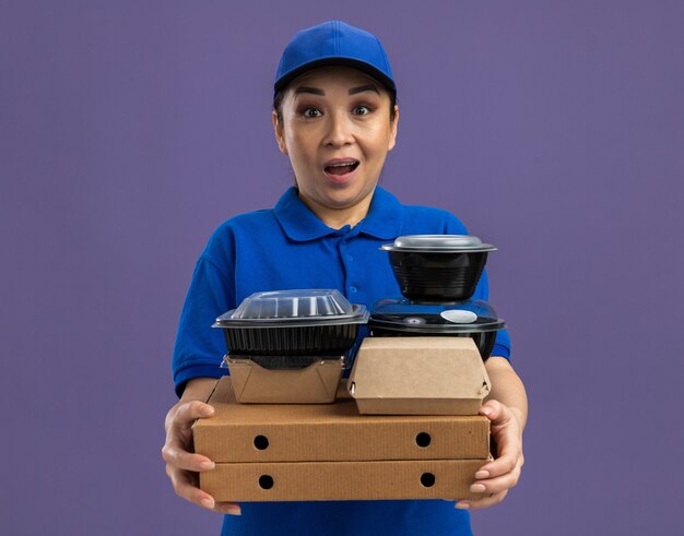 Repartidor joven feliz y sorprendido en uniforme azul y gorra sosteniendo cajas de pizza y paquetes de alimentos de pie sobre la pared púrpura