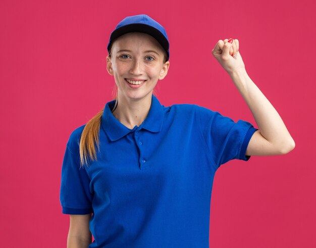 Repartidor joven feliz en uniforme azul y gorra sonriendo confiado levantando puño parado sobre pared rosa