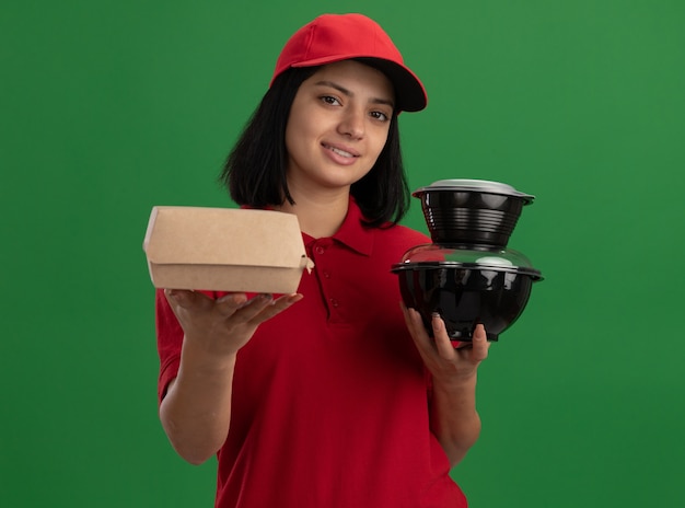 Foto gratuita repartidor joven feliz en uniforme rojo y gorra sosteniendo paquetes de alimentos mirando sonriente de pie amistoso sobre la pared verde