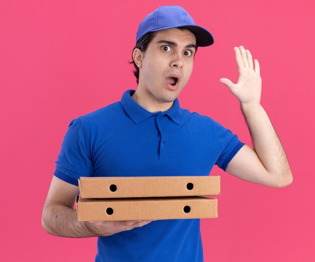 Repartidor joven impresionado con uniforme azul y gorra sosteniendo paquetes de pizza mirando al frente mostrando cinco con la mano aislada en la pared rosa