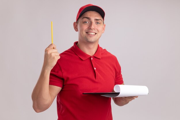 Repartidor joven impresionado con uniforme con gorra sosteniendo portapapeles levantando lápiz aislado en la pared blanca