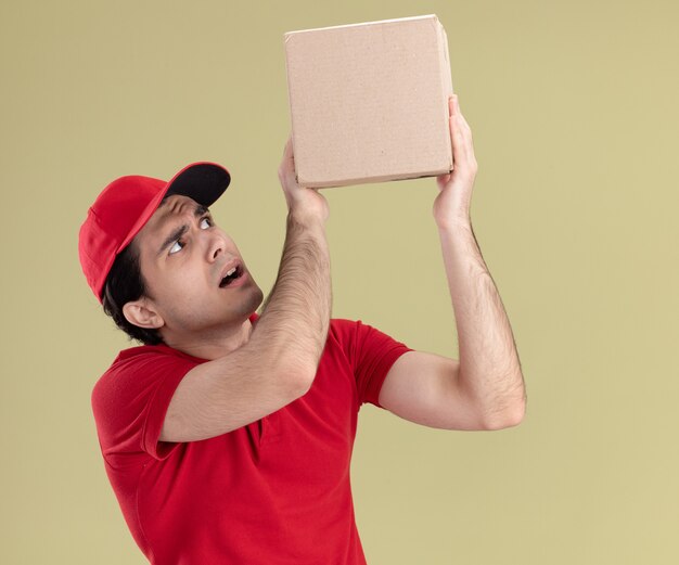 Repartidor joven impresionado en uniforme rojo y gorra levantando y mirando cardbox aislado en la pared verde oliva