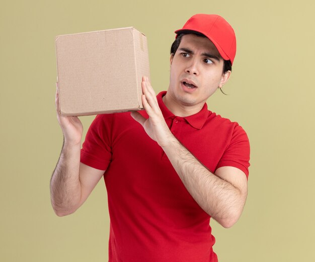 Repartidor joven impresionado en uniforme rojo y gorra sosteniendo y mirando cardbox aislado en la pared verde oliva