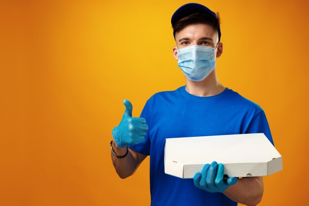 Repartidor joven en mascarilla y guantes con paquete de entrega de alimentos contra el fondo amarillo