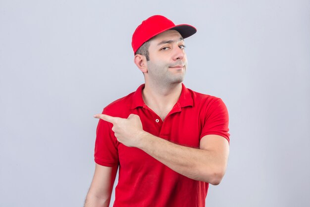 Repartidor joven en polo rojo y gorra apuntando hacia el lado con el dedo índice convencido y confiado de pie sobre fondo blanco aislado