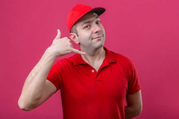 Repartidor joven en polo rojo y gorra haciendo gesto de llamarme que parece convencido y confiado sobre fondo rosa aislado