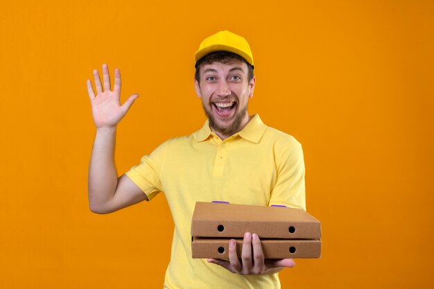 Repartidor joven positivo en camisa polo amarilla y gorra sosteniendo cajas de pizza mirando alegre haciendo gesto de bienvenida saludando con la mano en naranja aislado