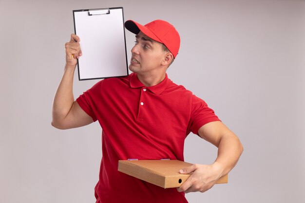 Repartidor joven preocupado vestido con uniforme con gorra sosteniendo la caja de pizza y mirando el portapapeles en su mano aislado en la pared blanca