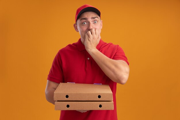 Repartidor joven preocupado vestido con uniforme con gorra sosteniendo cajas de pizza poniendo la mano en la boca aislada en la pared naranja