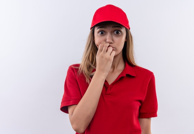 Repartidor joven preocupado vistiendo uniforme rojo y gorra poniendo la mano en la boca aislada en la pared blanca