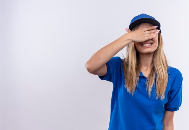 Repartidor joven sonriente con uniforme azul y gorra cubrió los ojos con la mano aislada en la pared blanca con espacio de copia