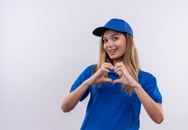 Repartidor joven sonriente con uniforme azul y gorra que muestra el gesto del corazón aislado en la pared blanca con espacio de copia