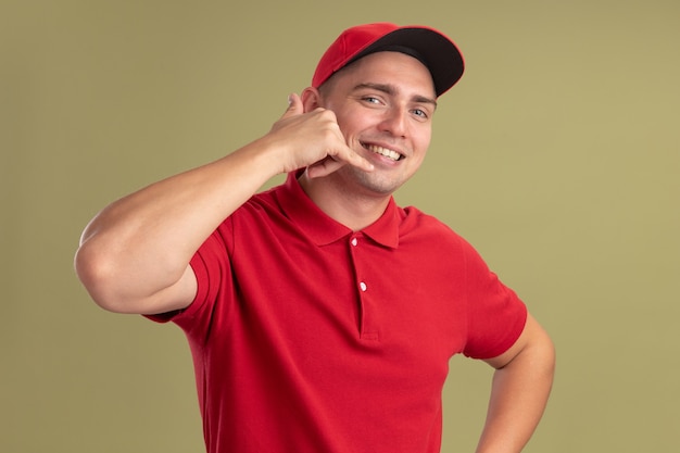 Repartidor joven sonriente con uniforme y gorra mostrando gesto de llamada telefónica aislado en la pared verde oliva