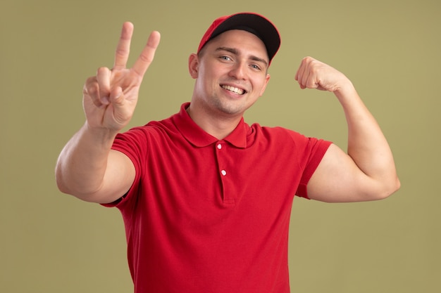 Repartidor joven sonriente con uniforme y gorra mostrando gesto de paz con gesto fuerte aislado en la pared verde oliva