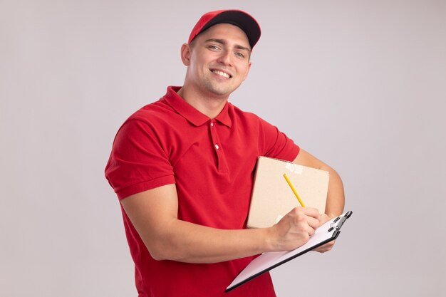 Repartidor joven sonriente con uniforme con gorra sosteniendo la caja y escribiendo algo en el portapapeles aislado en la pared blanca