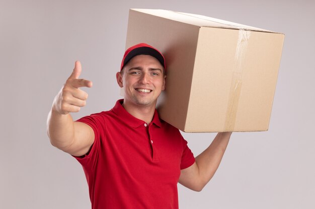 Repartidor joven sonriente con uniforme con gorra sosteniendo una caja grande en el hombro y apunta a la cámara aislada en la pared blanca