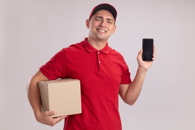 Repartidor joven sonriente con uniforme con gorra sosteniendo la caja y el teléfono aislado en la pared blanca