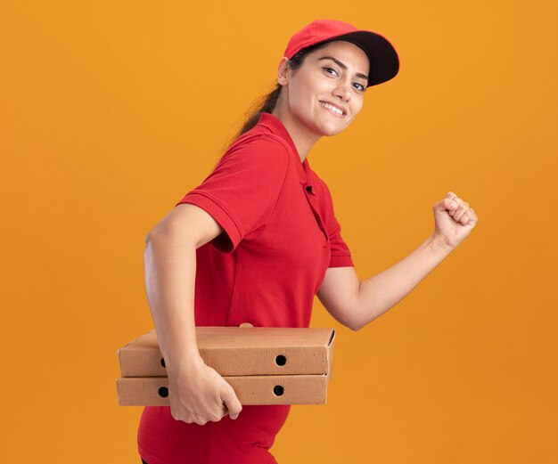 Repartidor joven sonriente con uniforme y gorra sosteniendo cajas de pizza mostrando gesto de ejecución aislado en la pared naranja