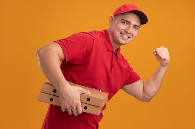 Repartidor joven sonriente con uniforme con gorra sosteniendo cajas de pizza mostrando un gesto fuerte aislado en la pared naranja