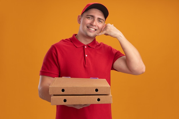 Repartidor joven sonriente con uniforme con gorra sosteniendo cajas de pizza mostrando gesto de llamada telefónica aislado en la pared naranja