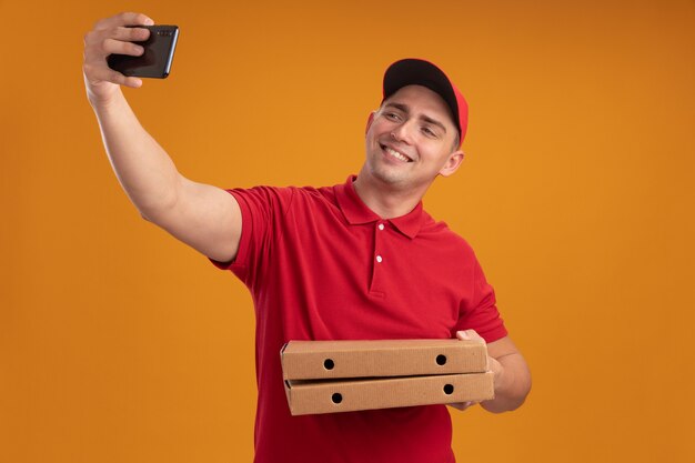 Repartidor joven sonriente con uniforme con gorra sosteniendo cajas de pizza tomar un selfie aislado en la pared naranja