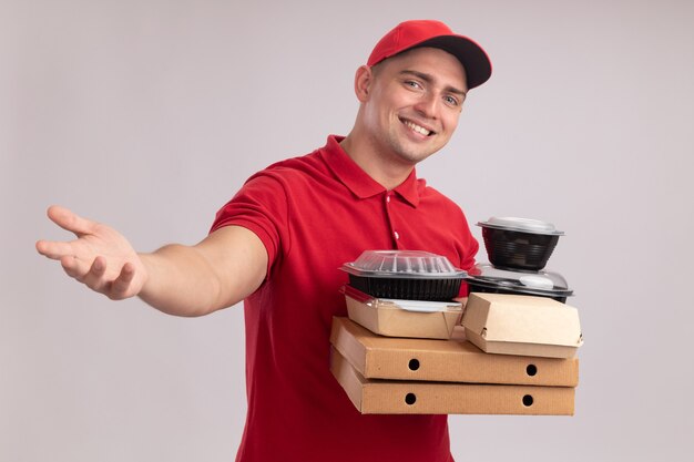 Repartidor joven sonriente con uniforme con gorra sosteniendo contenedores de comida en cajas de pizza sosteniendo la mano a la cámara aislada en la pared blanca