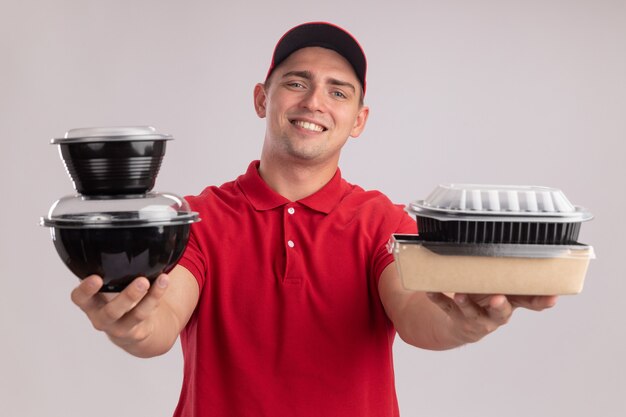 Repartidor joven sonriente con uniforme con gorra sosteniendo contenedores de comida en la cámara aislada en la pared blanca