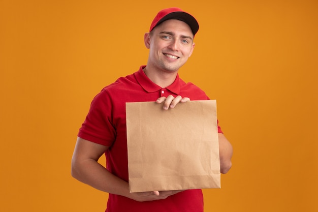 Repartidor joven sonriente con uniforme con gorra sosteniendo el paquete de alimentos de papel aislado en la pared naranja