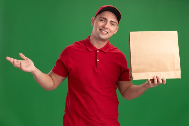 Repartidor joven sonriente con uniforme y gorra sosteniendo el paquete de alimentos de papel y extendiendo la mano aislada en la pared verde