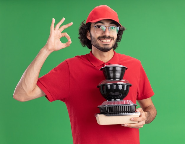 Repartidor joven sonriente en uniforme rojo y gorra con gafas sosteniendo paquetes de alimentos de papel y contenedores de alimentos mirando al frente haciendo bien signo aislado en la pared verde